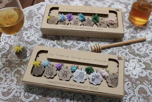 Two boxes of wooden honey dipper sets on a lace tablecloth with a glass of honey and dipper. Tea sachets included with set.