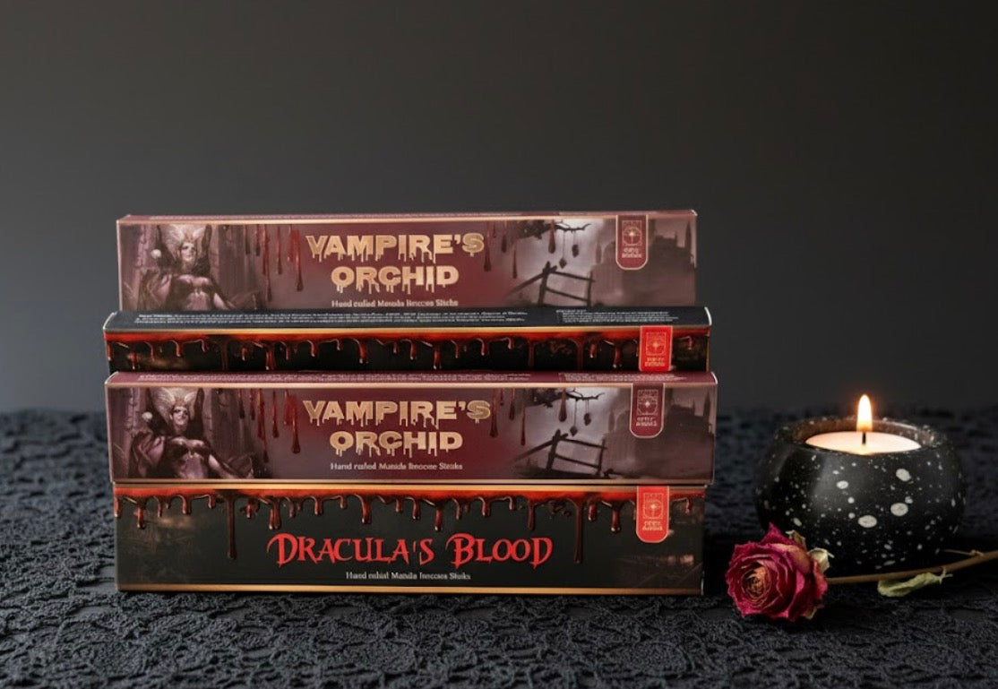 Stack of 'Vampire's Orchid' and 'Dracula's Blood' incense boxes with a candle and rose on a dark surface.