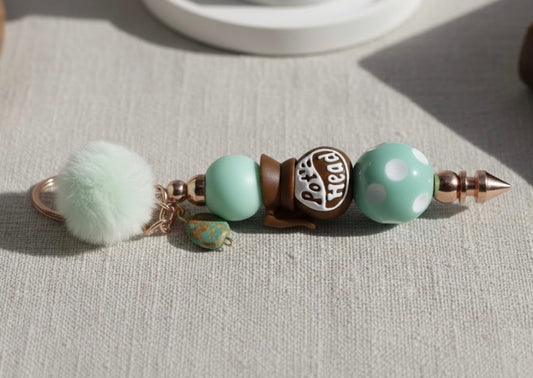 Coffee Pot Head Keychain with pom-pom and beads on a light surface