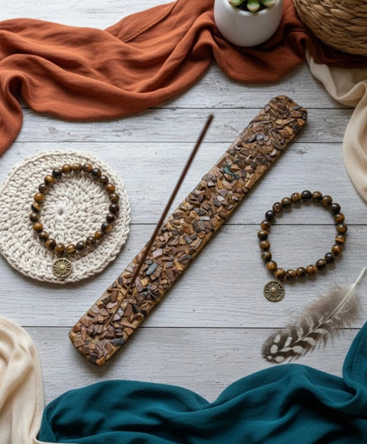 Decorative items including a tigers eye + wooden incense holder, beads, and a feather on a textured surface with fabric.