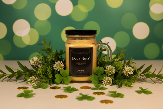 Jar of 'Deez Nutz' candle with decorative greenery and gold coins on a white surface with a bokeh green background.