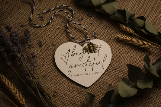 Heart-shaped tag with antique bronze honey bee charm 'beyond grateful' text on a textured surface with foliage.