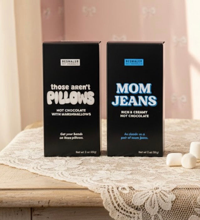 Two boxes of hot chocolate with marshmallows with fun sayings on a lace tablecloth.