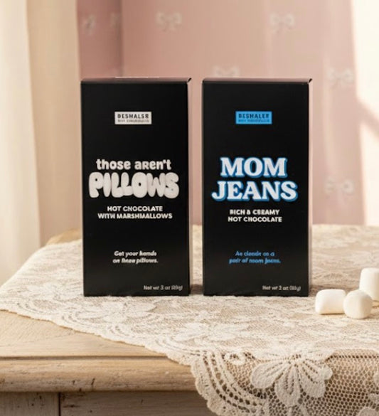 Two boxes of hot chocolate with marshmallows with fun sayings on a lace tablecloth.