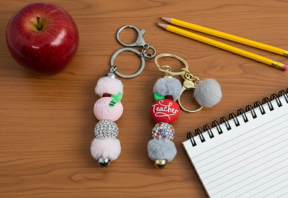 Two keychains for Teacher Appreciation with pom-poms on a wooden surface with an apple, pencils, and a notebook.