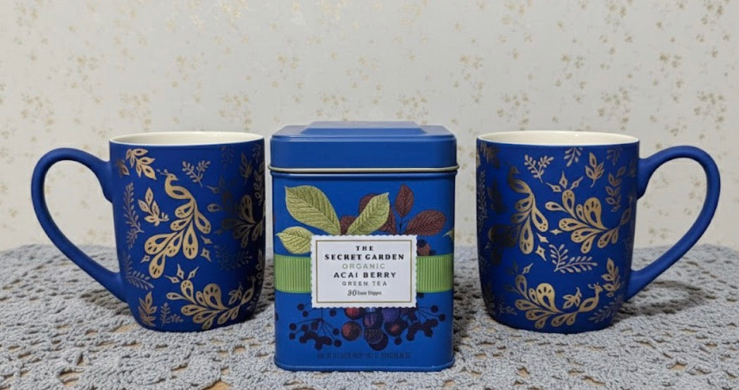 Two Matte blue peacock mugs with gold floral patterns and a tea tin labeled 'The Secret Garden' on a textured surface.