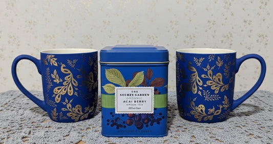 Two Matte blue peacock mugs with gold floral patterns and a tea tin labeled 'The Secret Garden' on a textured surface.