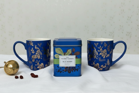Two matte blue mugs with gold leaf patterns and peacocks and a tea canister on a white surface.