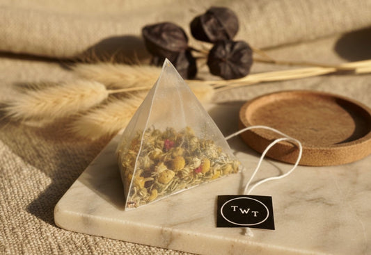 Pyramid tea bag with dried herbs on a marble surface, surrounded by dried plants and a wooden coaster.