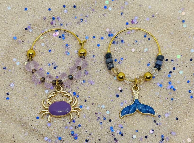 Two gold ocean wine glass charms with crab and mermaid tail charms on a glittery beige background. Purple and blue charms.