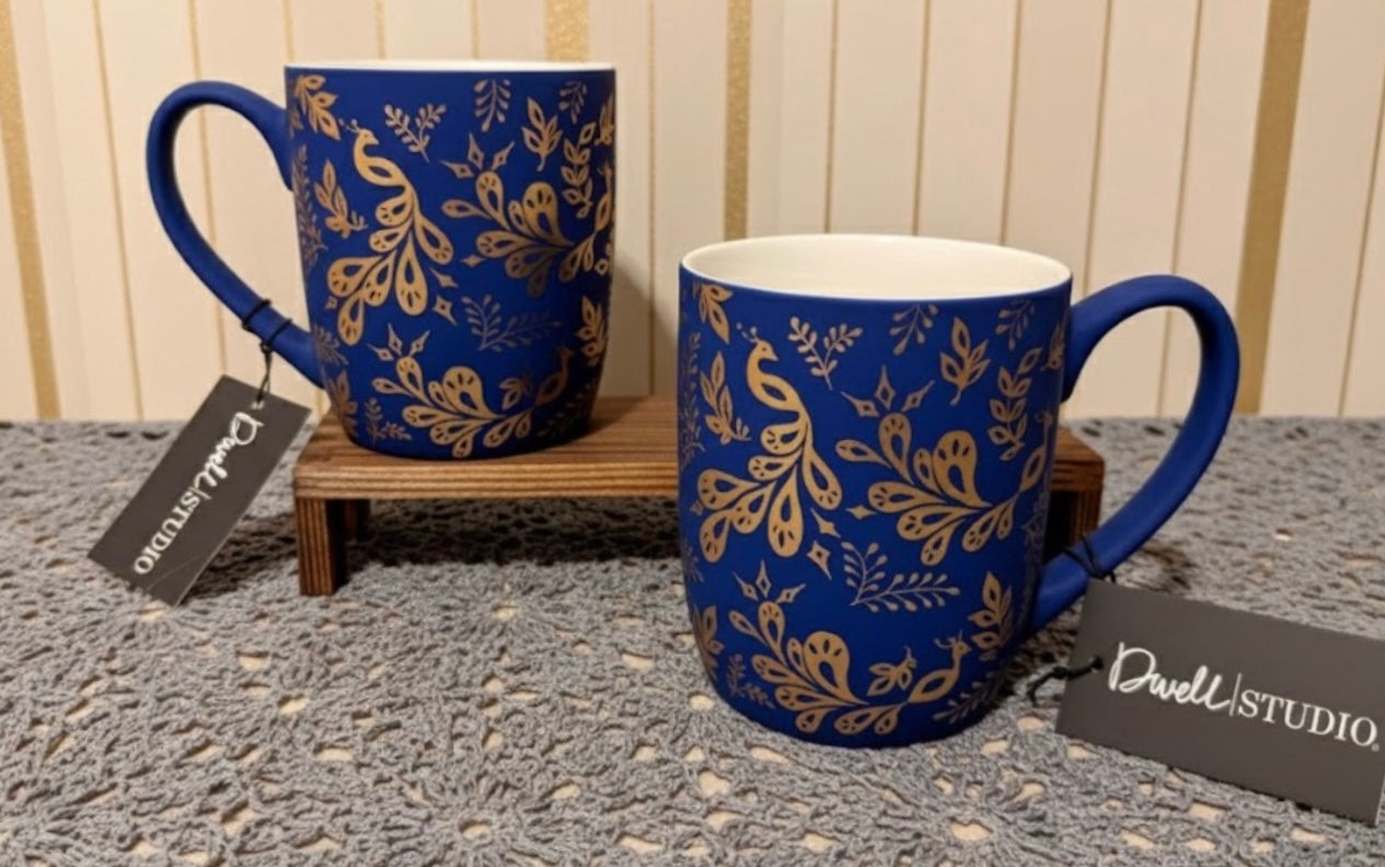 Two blue mugs with gold peacock patterns on a textured surface, featuring Dwell Studio tags.