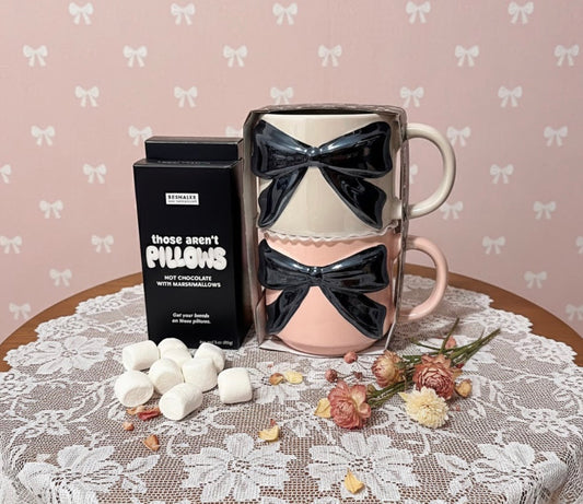 Two pink mugs with black bows, a box of hot chocolate mix, and marshmallows on a lace tablecloth.